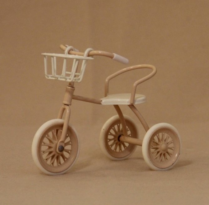 Beżowy miniaturowy rowerek trójkołowy z białym koszykiem na brązowym tle